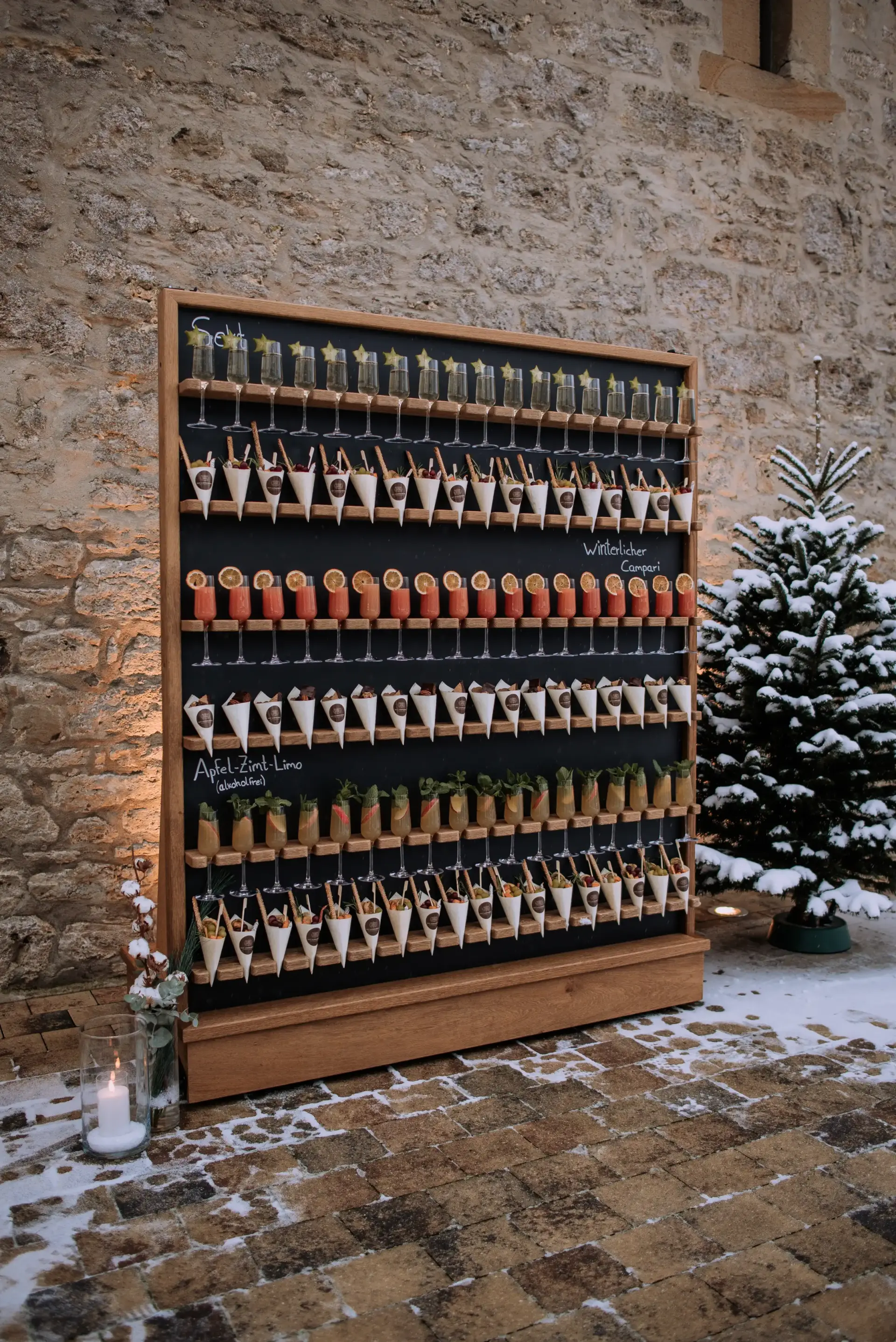 Sektwand mit winterlichen Getränken und Snacks auf einer Winterhochzeit. Daneben ein Tannenbaum und Kerzen und leichter Schnee auf dem Boden.