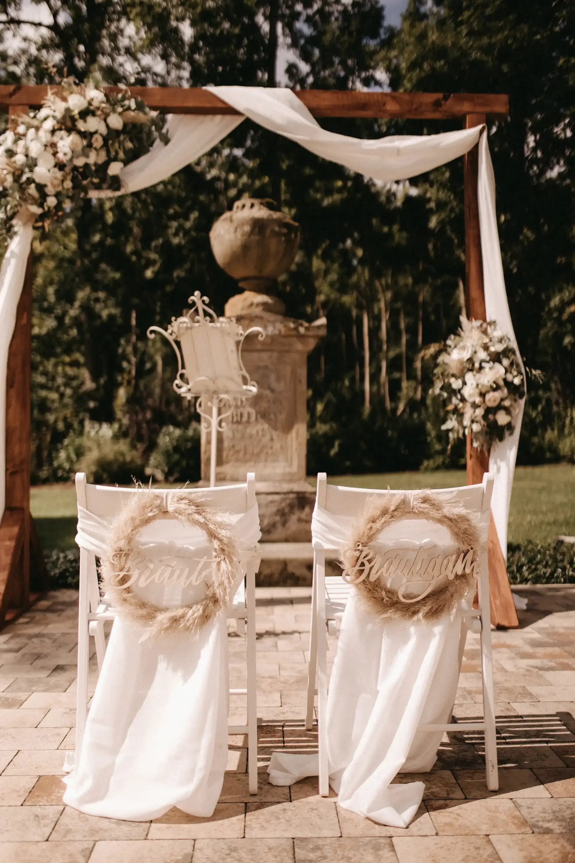 Hochzeits Setup im Eventgarten mit weißen Stühlen und 2 Kränzen mit der Beschriftung Braut und Bräutigam